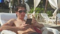 Young man wearing sunglasses using smartphone while lying on sunbed on tropical beach and drinking coconut cocktail Royalty Free Stock Photo