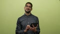 Young man using tablet over green background, wearing glasses, looking focused while holding device with both hands, presenting Royalty Free Stock Photo