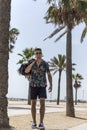 Young man walking on city wearing casual clothes and sunglasses with shorts flowered shirt holding travel bag Royalty Free Stock Photo