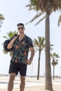 Young man walking on city wearing casual clothes and sunglasses with shorts flowered shirt holding travel bag Royalty Free Stock Photo