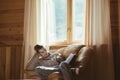 Young man in warm sweater reading by the window inside cozy log cabin