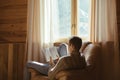 Young man in warm sweater reading by the window inside cozy log cabin