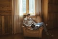 Young man in warm sweater reading by the window inside cozy log cabin