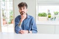 Young man wearing casual shirt sitting on white table with hand on chin thinking about question, pensive expression Royalty Free Stock Photo