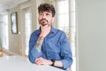 Young man wearing casual shirt sitting on white table with hand on chin thinking about question, pensive expression Royalty Free Stock Photo