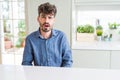 Young man wearing casual shirt sitting on white table In shock face, looking skeptical and sarcastic, surprised with open mouth Royalty Free Stock Photo