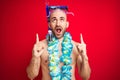 Young man wearing diving snorkel goggles and hawaiian lei flowers over isolated red background amazed and surprised looking up and