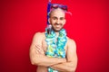 Young man wearing diving snorkel goggles and hawaiian lei flowers over isolated red background happy face smiling with crossed