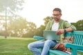 Young man wearing eyeglasses looking focused while working using laptop, sitting on the bench in the green park Royalty Free Stock Photo