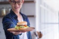 Young man wearing glasses smiling, looking at the camera, and holding a homemade hamburger on a wooden dish with blur background. Royalty Free Stock Photo