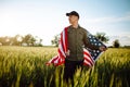 Young man wearing green shirt and cap stands wrapped in the american flag at the green wheat field. Patriotic boy celebrates usa Royalty Free Stock Photo