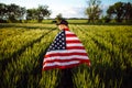 Young man wearing green shirt and cap stands wrapped in the american flag at the green wheat field. Patriotic boy celebrates usa Royalty Free Stock Photo