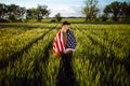 Young man wearing green shirt and cap stands wrapped in the american flag at the green wheat field. Patriotic boy celebrates usa Royalty Free Stock Photo