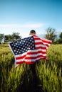 Young man wearing green shirt and cap stands wrapped in the american flag at the green wheat field. Patriotic boy celebrates usa Royalty Free Stock Photo