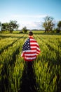 Young man wearing green shirt and cap stands wrapped in the american flag at the green wheat field. Patriotic boy celebrates usa Royalty Free Stock Photo