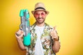 Young man wearing hawaiian flowers shirt holding water gun over yellow isolated background happy with big smile doing ok sign, Royalty Free Stock Photo