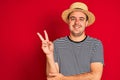 Young man wearing striped navy t-shirt and hat standing over isolated red background smiling with happy face winking at the camera Royalty Free Stock Photo