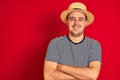 Young man wearing striped navy t-shirt and hat standing over  red background happy face smiling with crossed arms looking Royalty Free Stock Photo