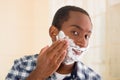 Young man wearing white blue square pattern shirt applying shaving foam onto face using hands, looking into camera Royalty Free Stock Photo