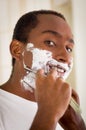 Young man wearing white t-shirt shaving using shavette, foam on face, looking into camera Royalty Free Stock Photo