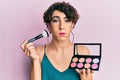 Young man wearing woman make up holding makeup brush and blush puffing cheeks with funny face