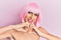 Young man wearing woman make up wearing pink wig smiling in love doing heart symbol shape with hands Royalty Free Stock Photo