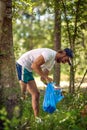 Young man in white shirt in a green spring forest in a sunny weather, holding blue plastic bag collecting trash. Volunteers in Royalty Free Stock Photo
