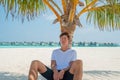Young man in white t-shirt and black shorts relaxing under palm tree at tropical sandy beach at island luxury resort Royalty Free Stock Photo