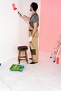 young man in working overall and headband painting wall in red by paint roller near chair Royalty Free Stock Photo