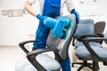 Young man in workwear and rubber gloves cleans the office chair with professional equipment.