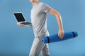 Young man with yoga mat and laptop running on color background. Concept of balance between rest and work