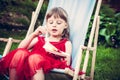 Young mannered girl in red dress relaxing in chaise lounge eat cake during outdoor party in the garden