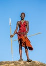 Young Masai warrior is standing on a big stone in traditional clothing with a spear in the savannah against a blue sky.