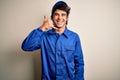 Young mechanic man wearing blue cap and uniform standing over isolated white background smiling doing phone gesture with hand and Royalty Free Stock Photo