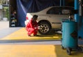 Young mechanic in a red coverall working on a car inside a workshop or garage. She is squatting beside the rear wheel of a car and Royalty Free Stock Photo