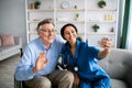 Young medical worker and her older handicapped patient taking selfie together indoors. Home care service for seniors Royalty Free Stock Photo