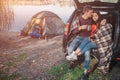 Young man embrace woman. They sit in trunk. Model is covered with blanket. Couple is at lake. There is tent at waterline.