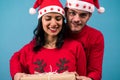 A young man in Santa`s hat staring at the camera next to his girlfriend, a brunette dressed in Christmas pajamas, holding a