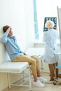 Young man sitting on a hospital daybed leaning on wall and touching his forehead with doctor examining his