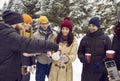Young men and women with hot tea in paper disposable cups walk through snowy forest on winter day.