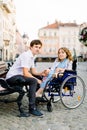 Young man and woman in wheelchair have fun and smiling on the bench outside. Old city street, love and disability Royalty Free Stock Photo