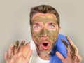 Young messy funny man looking at himself horrified in bathroom mirror with green cream on his face applying beauty facial mask pro