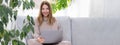 Young or middle age woman sitting with laptop on grey couch in home office with monstera plant. Concept of remote Royalty Free Stock Photo