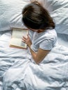 Young millennial candid woman is reading red book at home on white striped bedsheets. Early morning. Royalty Free Stock Photo