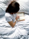 Young millennial candid woman reading red book at home on white striped bedsheets. Early morning. Text in book is Royalty Free Stock Photo
