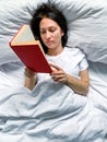 Young millennial candid woman is reading red book at home on white striped bedsheets. Royalty Free Stock Photo