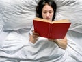 Young millennial candid woman is reading red book at home on white striped bedsheets. Royalty Free Stock Photo