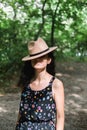 Young millennial woman in dress and beige hat stands in forest happy and relaxed, enjoys nature and tranquility. Travel