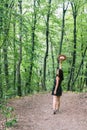 Young millennial woman in dress and beige hat stands in forest happy and relaxed, enjoys nature and tranquility. Travel