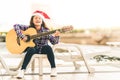 Young mixed race girl playing guitar, singing and smiling joyfully by swimming pool, with christmas santa hat